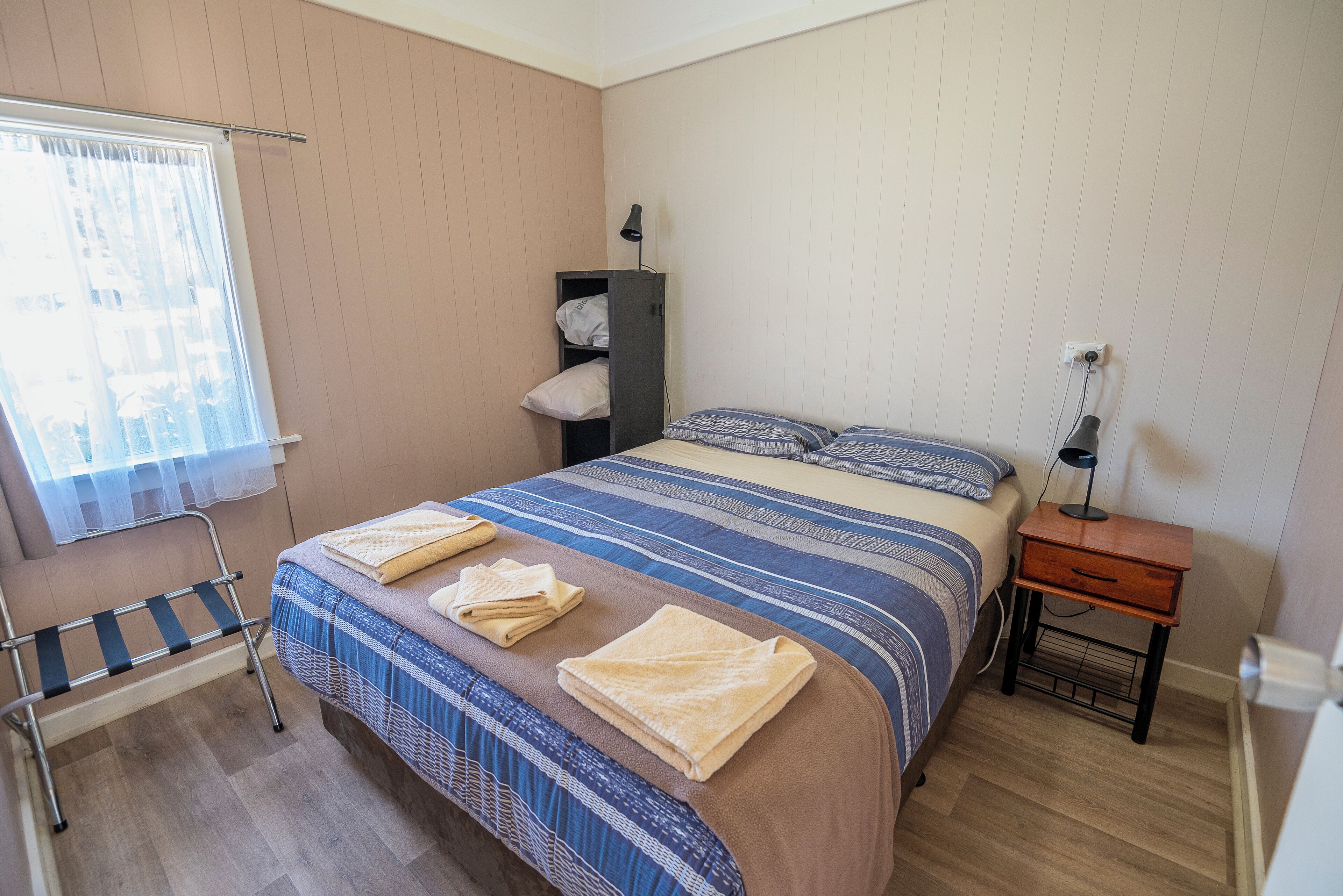 Una cama bien hecha con ropa de cama a rayas y dos toallas, una mesita de noche con lámpara, una silla y un armario junto a la pared.