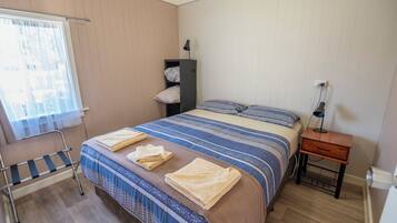 Una cama bien hecha con ropa de cama a rayas y dos toallas, una mesita de noche con lámpara, una silla y un armario junto a la pared.