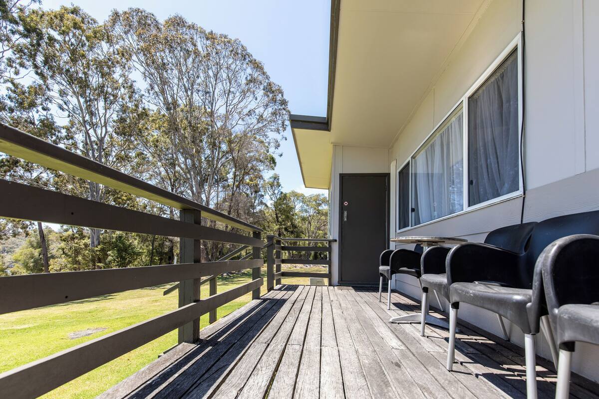 A wooden deck with a railing, a row of black chairs, and a building with large windows.