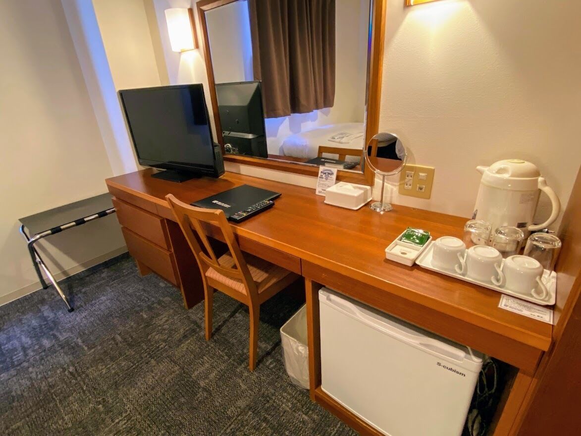 Una habitación de hotel con un escritorio de madera, televisión, espejo, refrigerador pequeño y cafetera.