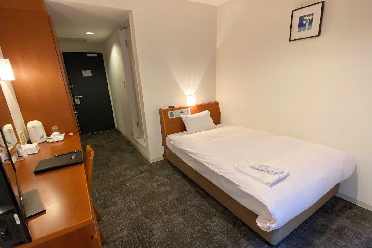 A hotel room with a single bed, a desk with a laptop, a chair, and a door leading to another room.