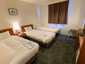 A hotel room with three beds, a desk, a chair, a lamp, and a window with curtains.