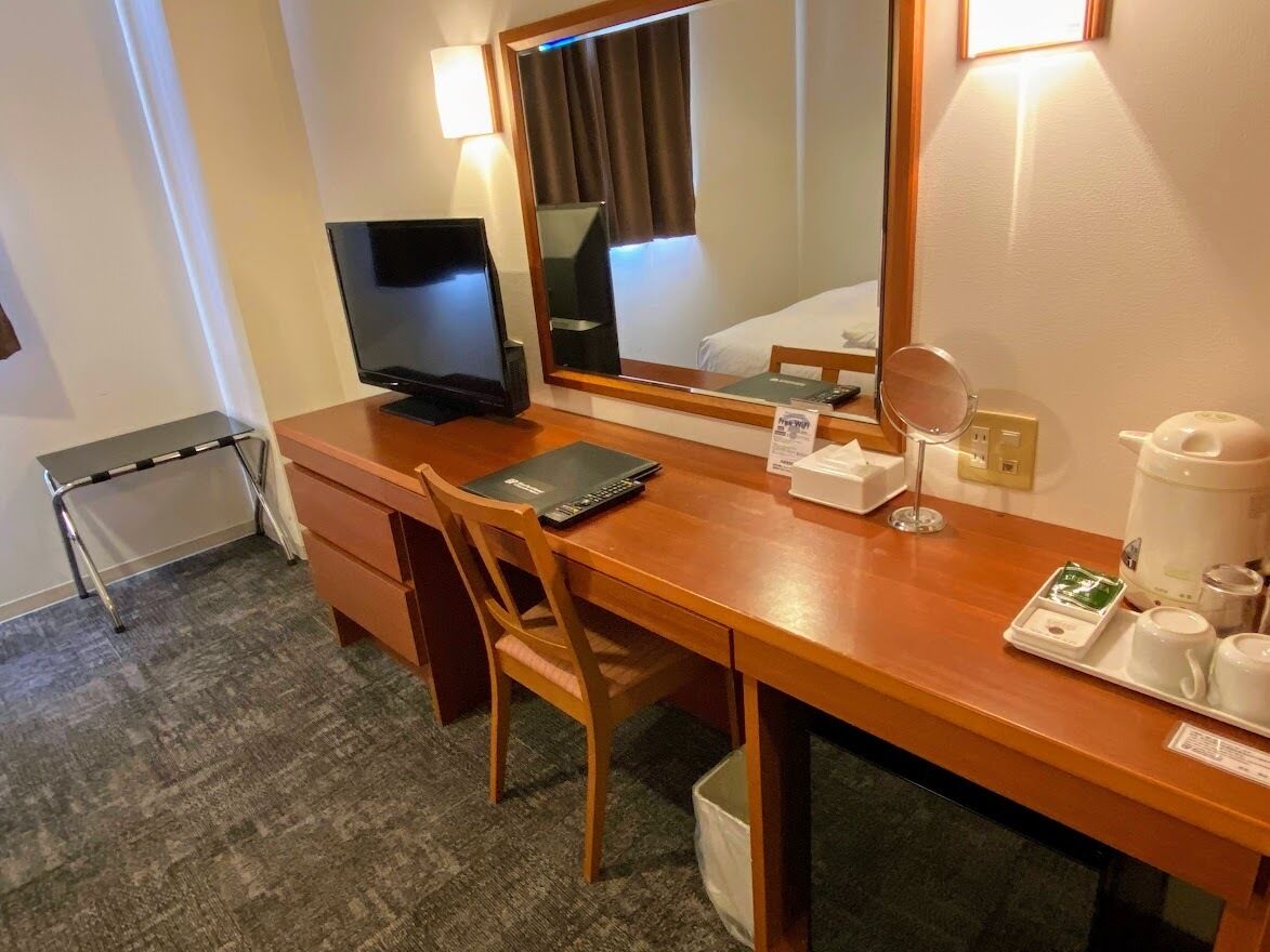 A hotel room with a wooden desk, a television, a mirror, a coffee maker, and a small tray with cups and a remote control.