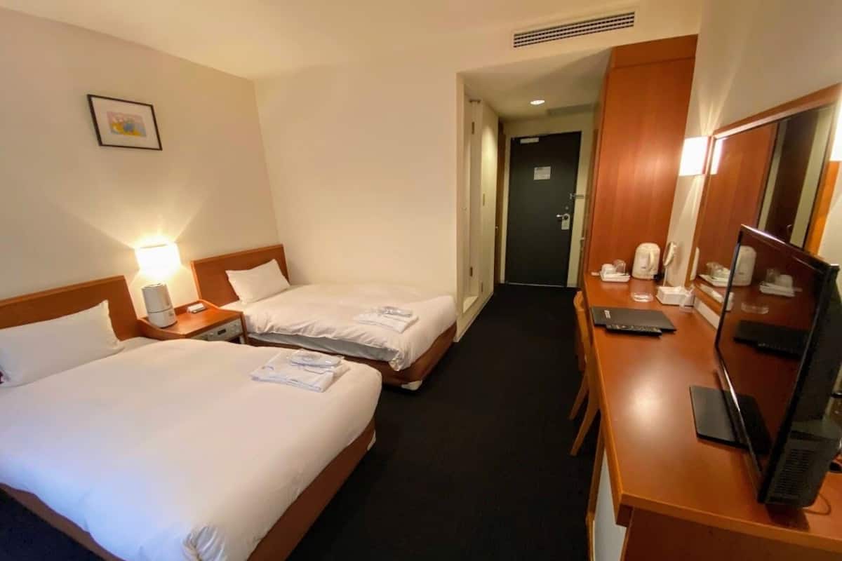 A hotel room with two beds, a desk with a computer, a mirror, and a door leading to another room.