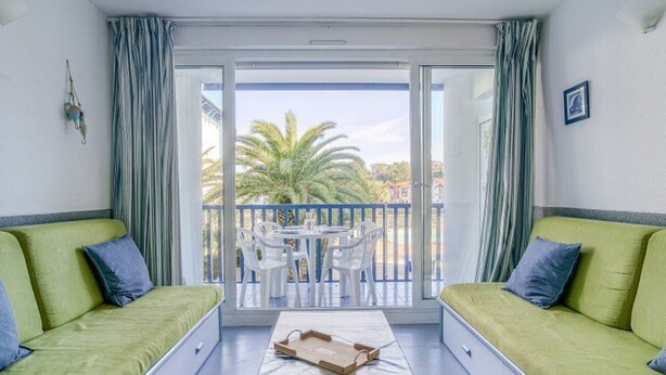 A room with two green sofas, a small table, and a balcony with a view of palm trees and buildings. - Beachside Apartment with Pool and Terrace in Urrugne (Urrugne)