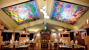A restaurant with high ceilings, large colorful murals, and a central light fixture. Tables are set with white napkins and silverware. - Toshali Sands Nature Escape (Puri)