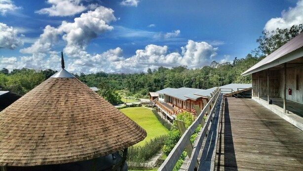 A wooden deck with a thatched roof structure, a building with a corrugated metal roof, and a view of a landscaped area with trees and a clear sky. - Borneo Tribal Village (Kuching)
