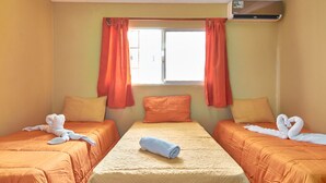 Quarto de hotel com duas camas, roupa de cama laranja, janela com cortinas e aparelho de ar condicionado.