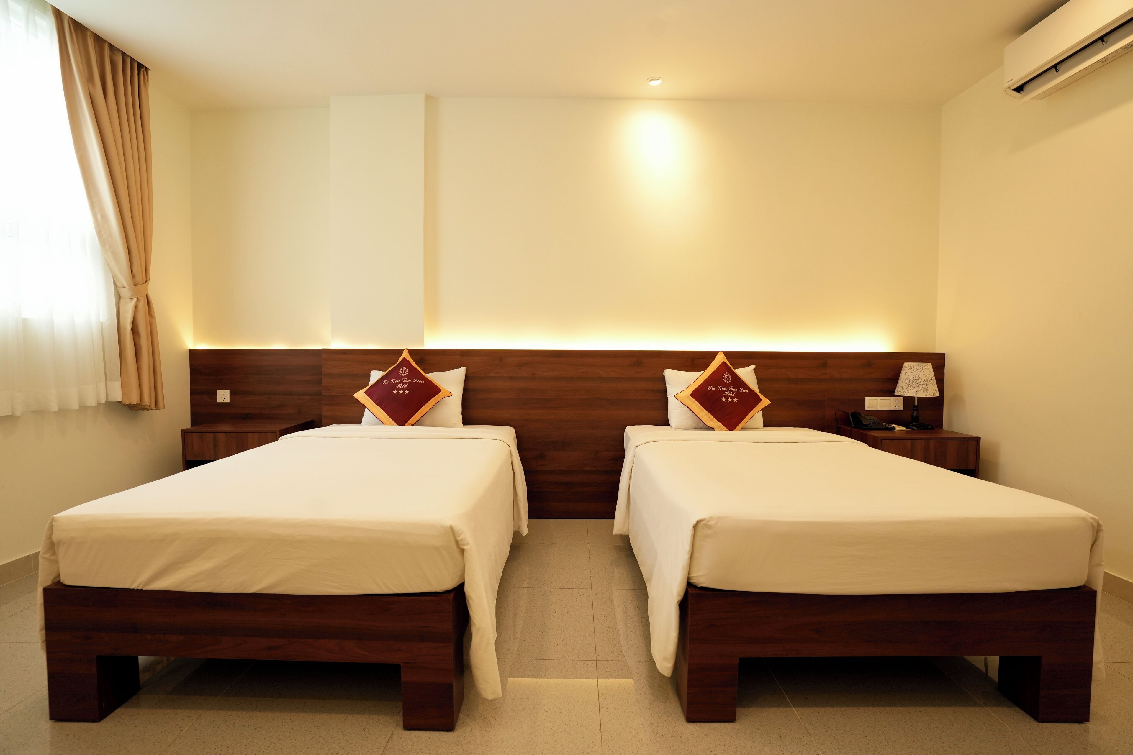 A hotel room with two single beds, a wooden headboard, a nightstand with a lamp, and a window with curtains.