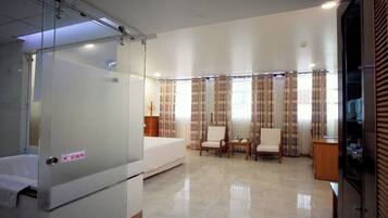 A hotel room with a bed, two chairs, a small table, and a glass partition.