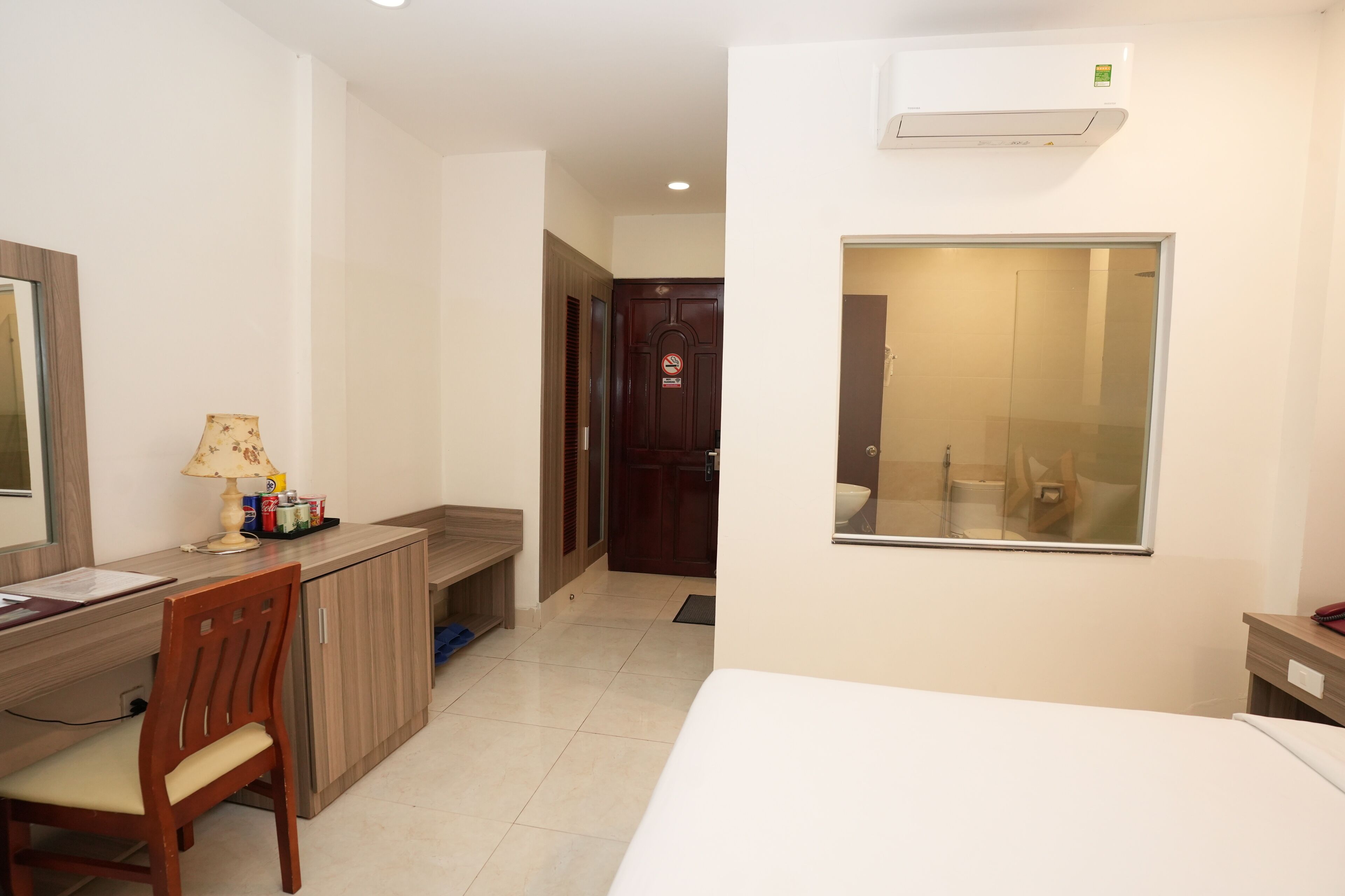 A hotel room with a bed, a desk with a chair, a lamp, a mirror, and a bathroom visible through a glass door.