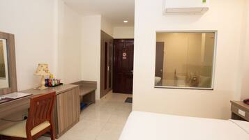 A hotel room with a bed, a desk with a chair, a lamp, a mirror, and a bathroom visible through a glass door.