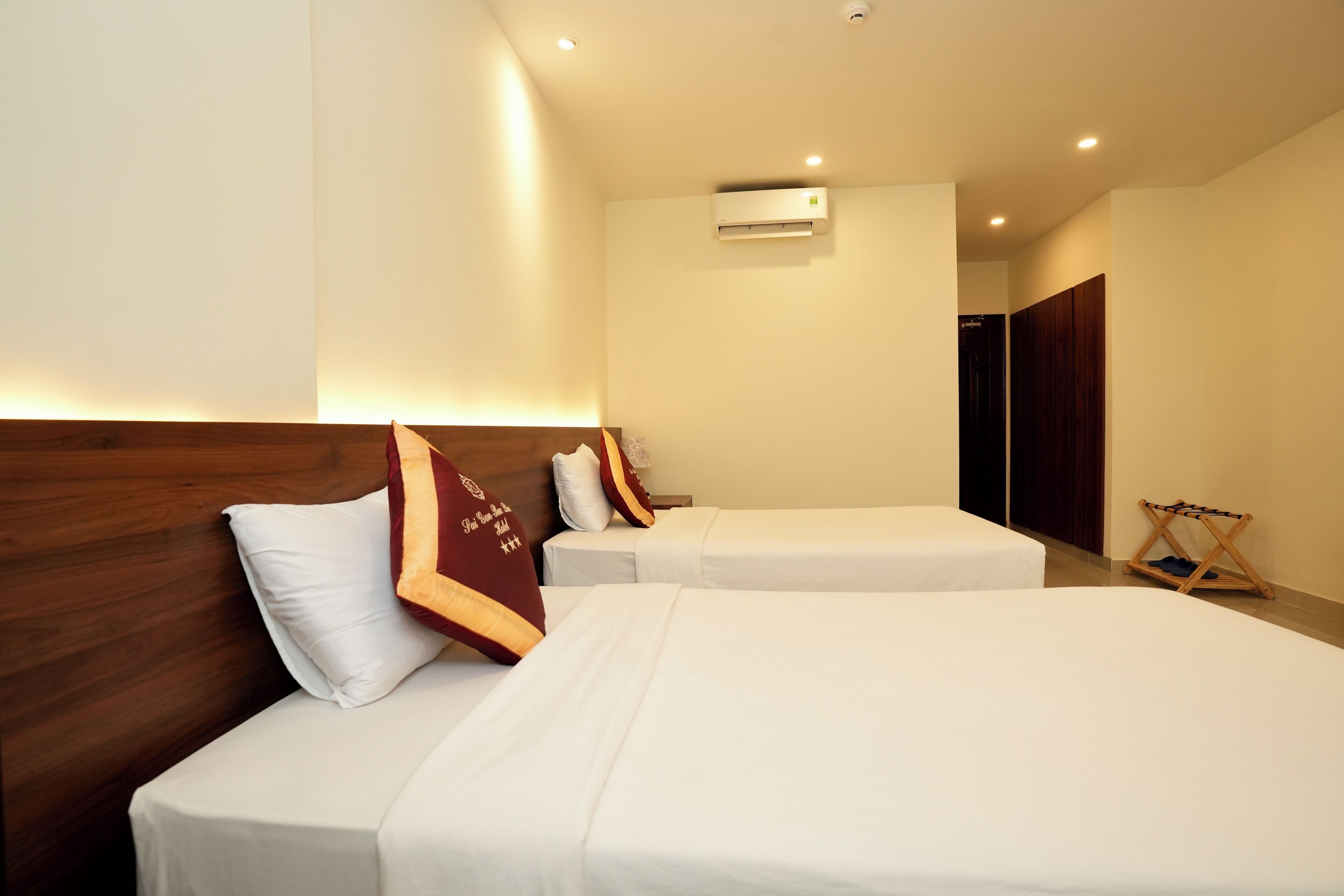 A hotel room with two single beds, a wooden headboard, a wall-mounted air conditioner, and a small wooden stool.