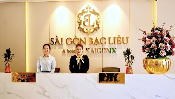 Een receptiebalie met een bord waarop staat 'SÀI GÒN BÀC LIÊU A hotel SAIGONX' en twee medewerkers die erachter staan.