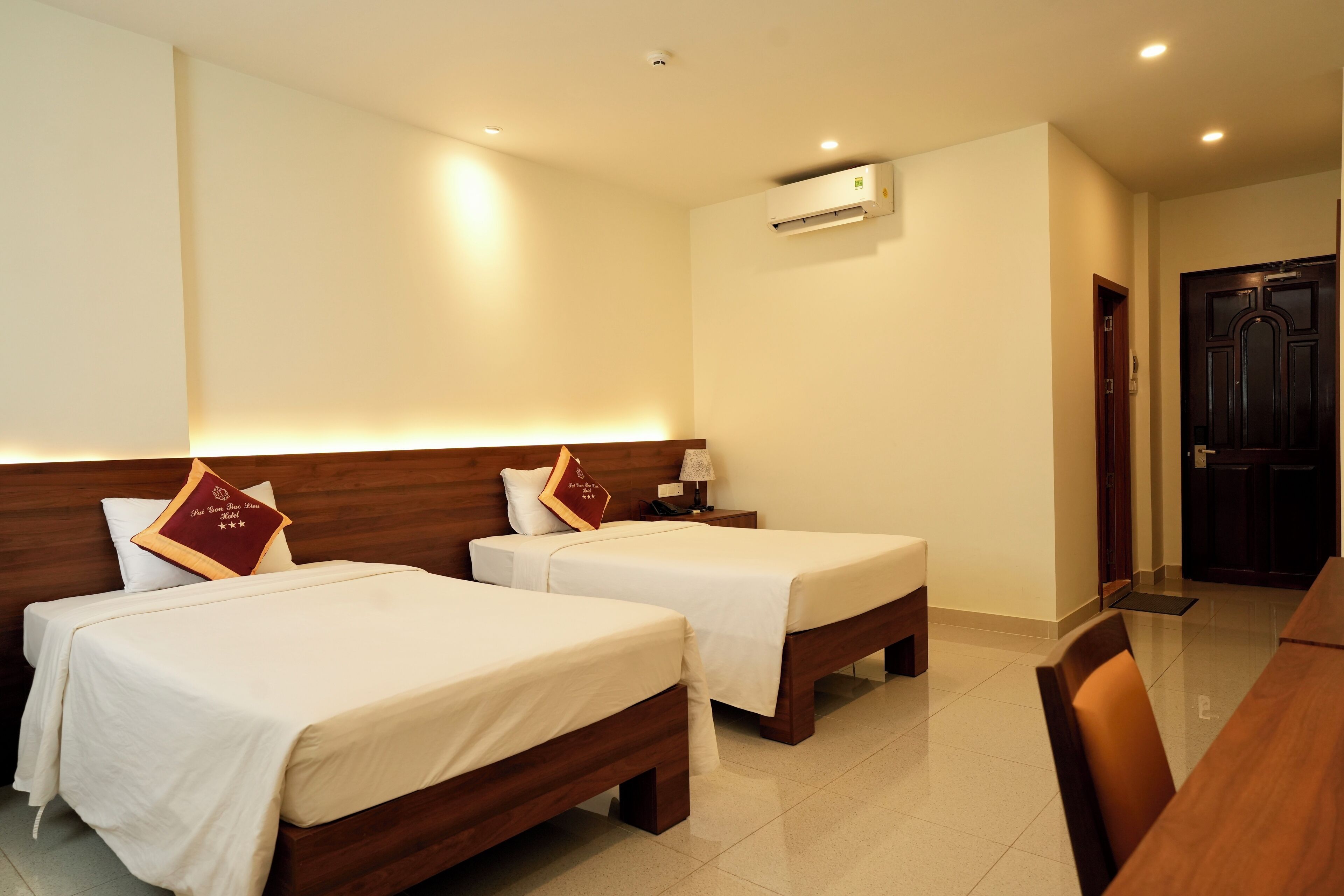 A hotel room with two beds, a wooden headboard, a bedside table with a lamp, and a door to the right.