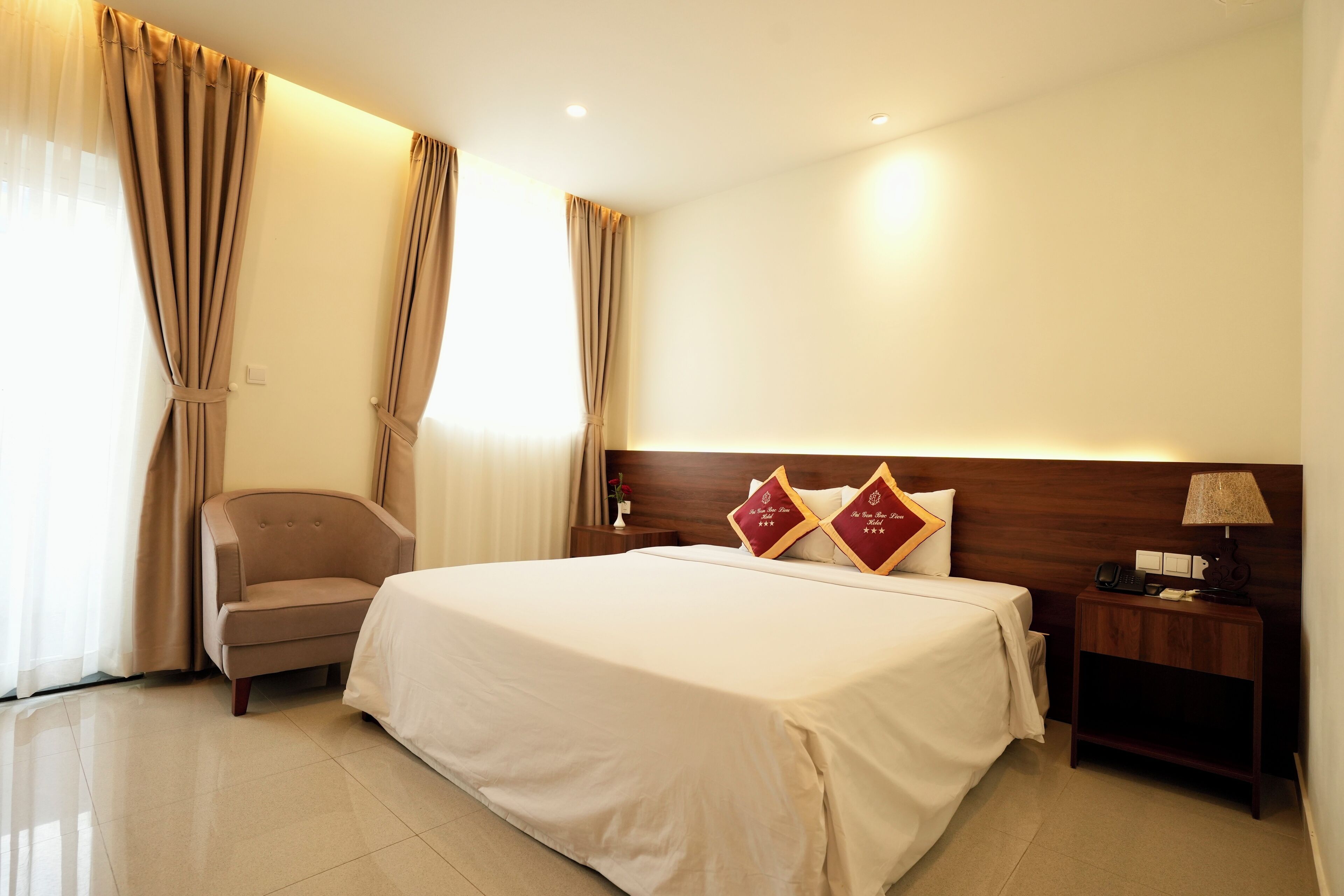 A hotel room with a large bed, a chair, a nightstand with a lamp, and curtains.