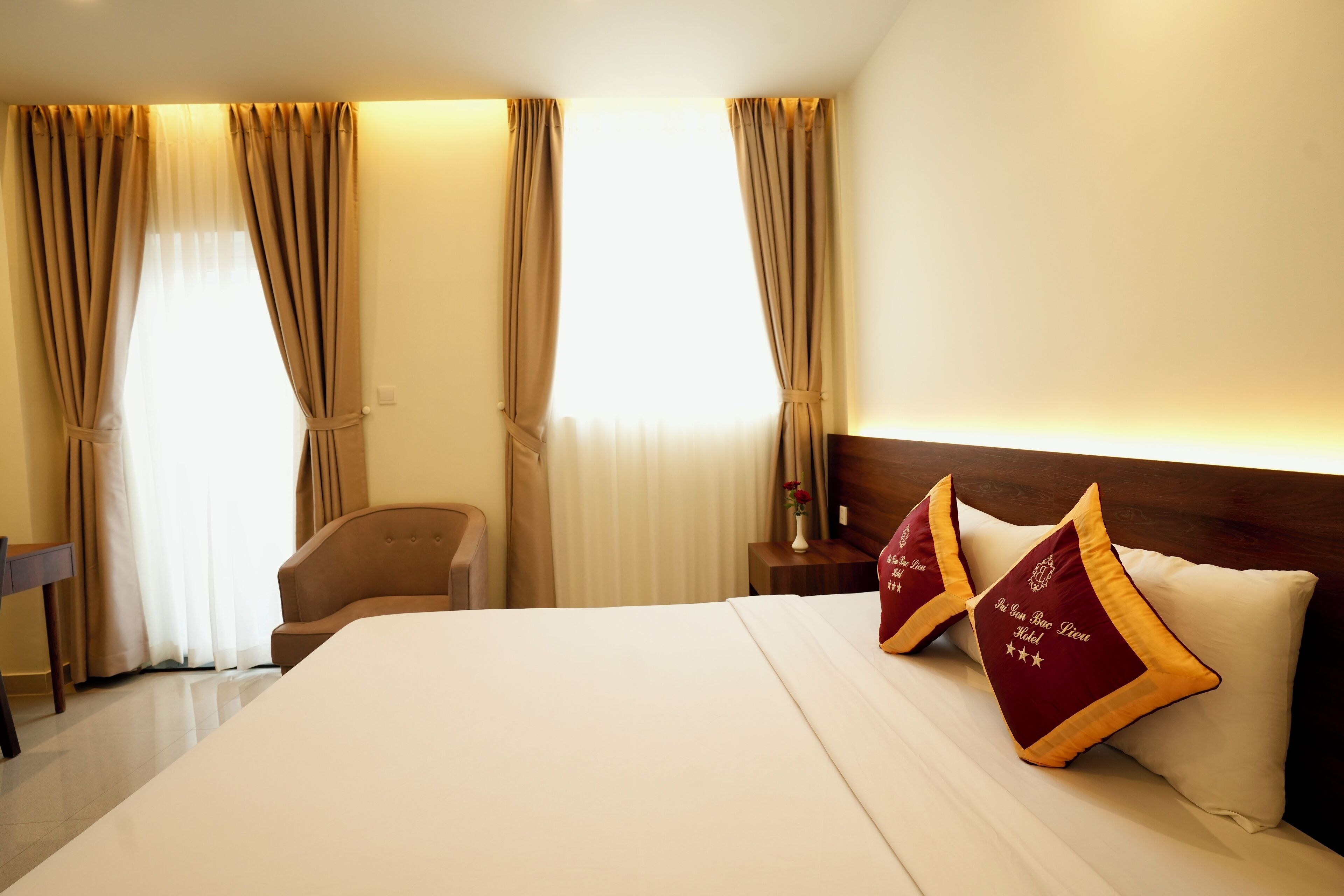 Hotelkamer met een groot bed, twee decoratieve kussens, een houten hoofdbord, een nachtkastje met een vaas en gordijnen.