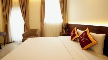 Hotelkamer met een groot bed, twee decoratieve kussens, een houten hoofdbord, een nachtkastje met een vaas en gordijnen.