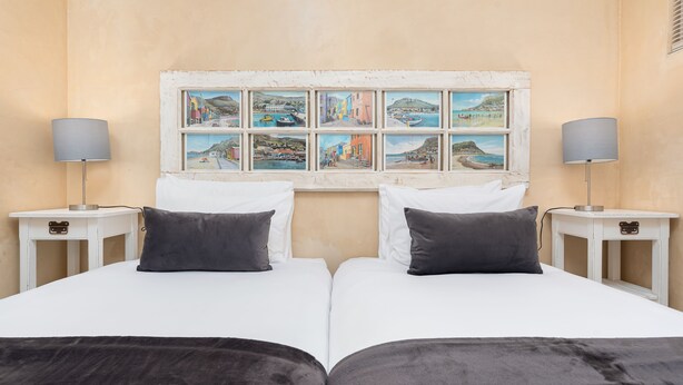 Uma cama de casal com lençóis brancos e duas almofadas escuras, com mesas de cabeceira com luminárias, num quarto com paredes bege e uma série de quadros costeiros emoldurados acima da cama.