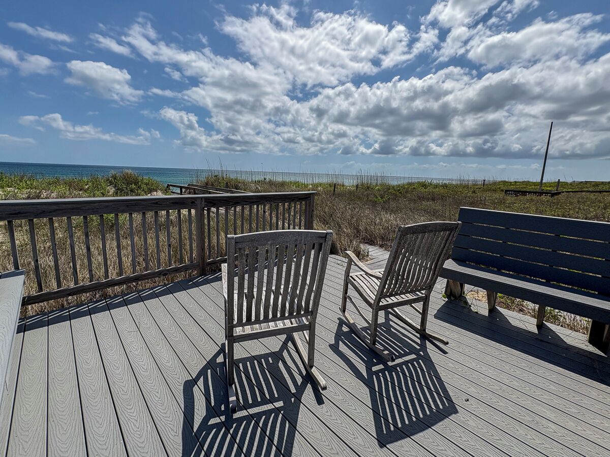 A wooden deck with two chairs and a bench, overlooking a beach with a fence and vegetation.