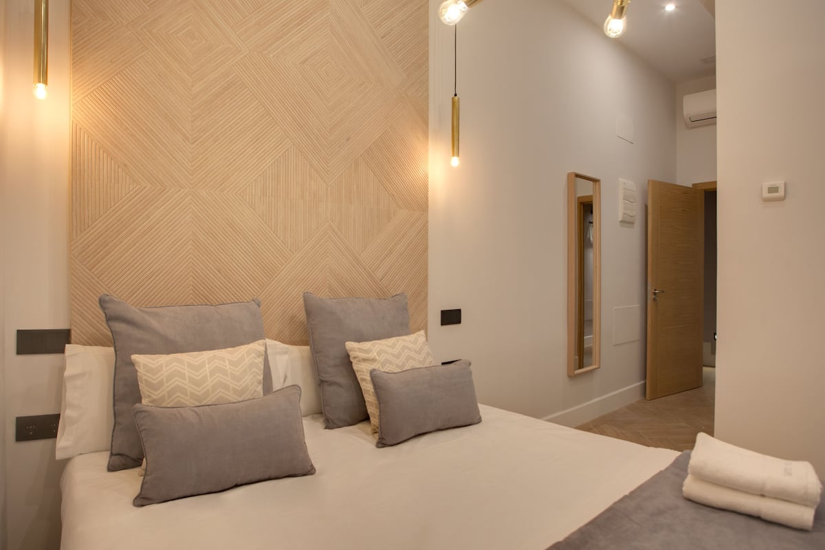 Un dormitorio moderno con una cama grande, varios almohadones, un cabecero de madera y una mesita de noche.