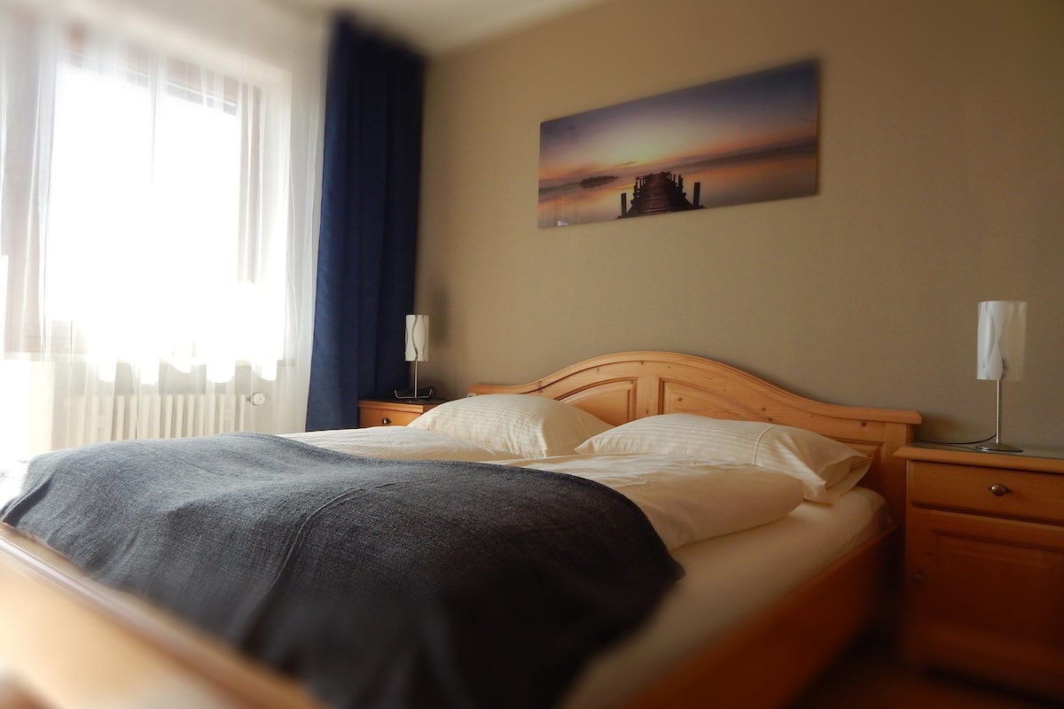 Una habitación de hotel con cama, mesita de noche, lámpara, un cuadro en la pared y una ventana con cortinas.