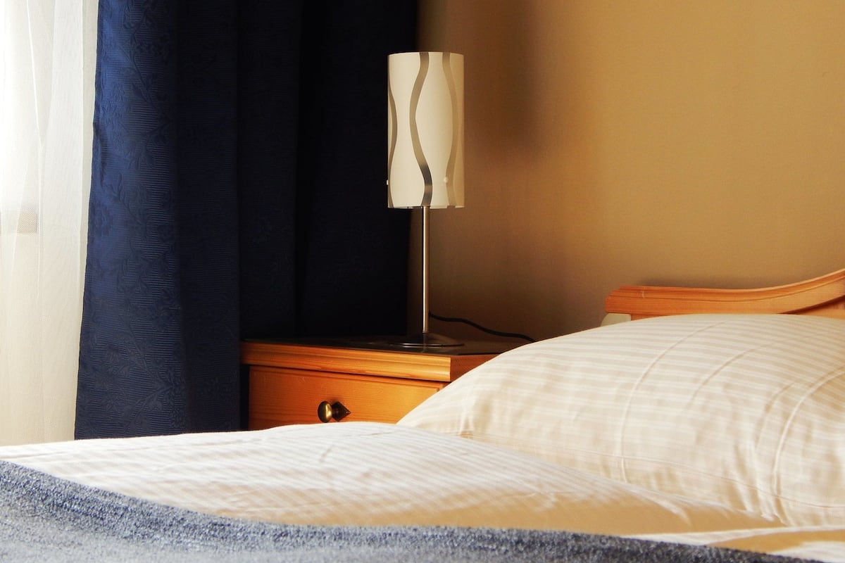 Una habitación de hotel con cama, mesita de noche, lámpara y cortina.