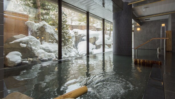 An indoor hot spring area with a wooden tub, rocks, and a view of a snowy outdoor scene through large windows.