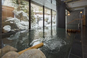 Ein Innenbereich mit Thermalquellen, einer Holzwanne, Felsen und Blick auf eine verschneite Landschaft durch große Fenster.