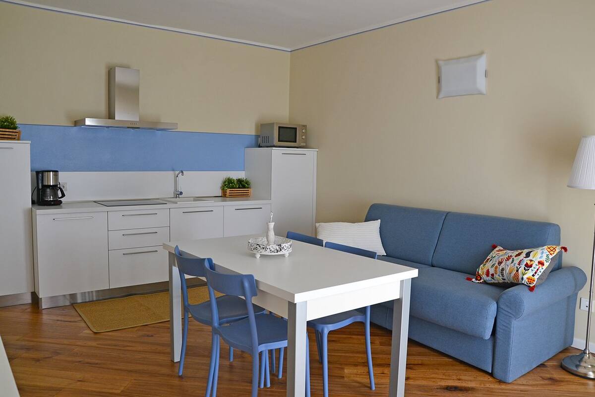 A compact living space with a kitchenette, a dining area with a white table and blue chairs, and a blue sofa with a patterned pillow.