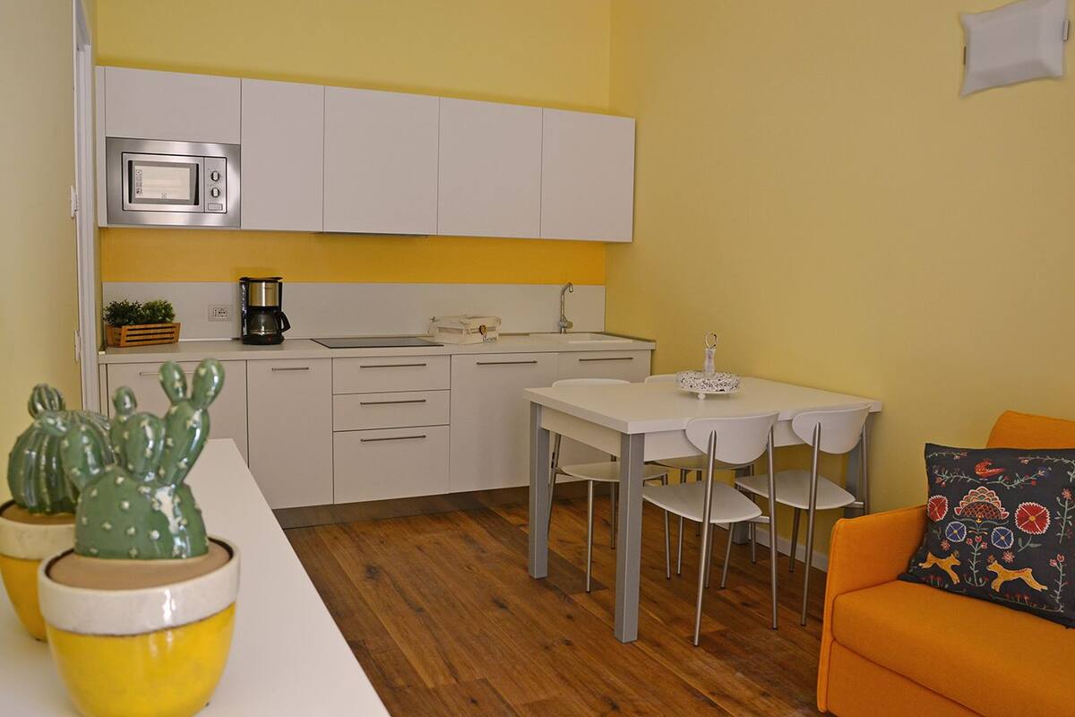 A compact kitchen with white cabinets, a microwave, a coffee maker, and a sink. There is a dining table with chairs and a small orange sofa with a patterned cushion.