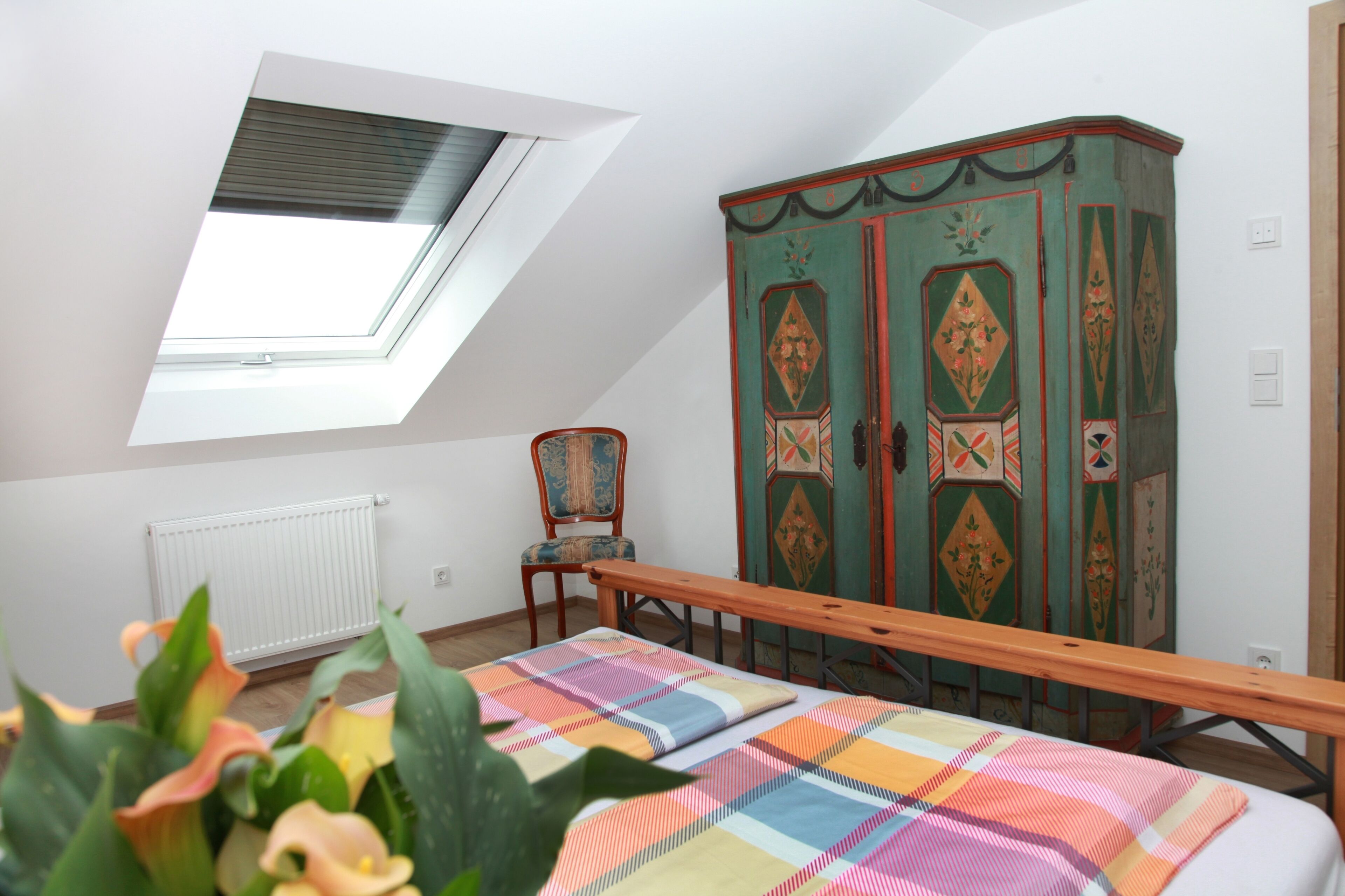 Een kamer met een schuine kap, een radiator, een houten bankje, een kleurrijke quilt, een groen kastje met bloemenmotieven, en een stoel.