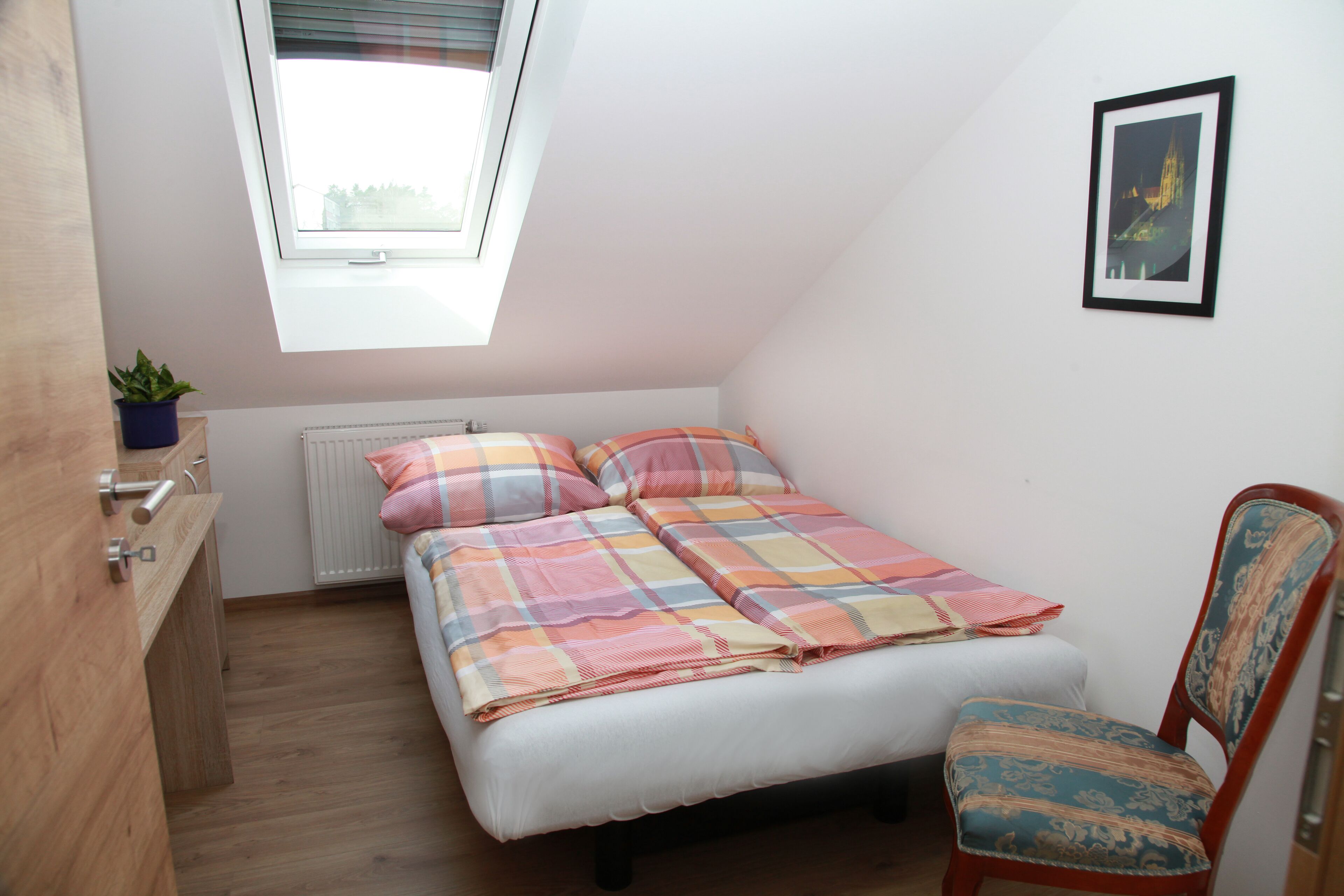 A small, well-lit bedroom with a bed, a chair, a radiator, a window, and a framed picture on the wall.