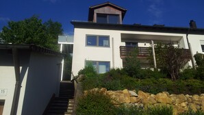 Moderni, kaksikerroksinen talo, jossa on parveke, näkyviin jätetty portaikko ja kivimuuri etualalla.