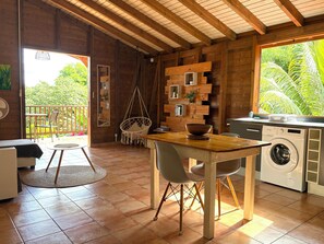 A wooden interior with a dining table, chairs, a hanging chair, a washing machine, and a view of greenery outside. - Bamboo Canyon Villa - Large F2 with Air Conditioning (Petit-Bourg)