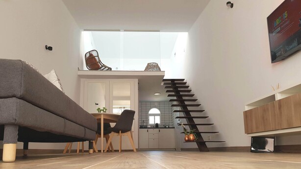 Ein modernes Wohnzimmer mit einer Couch, Sesseln, Holzboden, einer Treppe und einer Küche im Hintergrund.