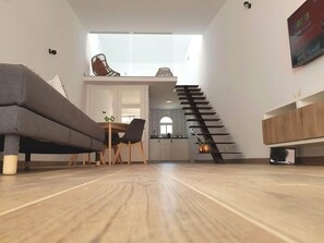 Un salon moderne avec un canapé, des fauteuils, un plancher en bois, un escalier et une cuisine en arrière-plan.