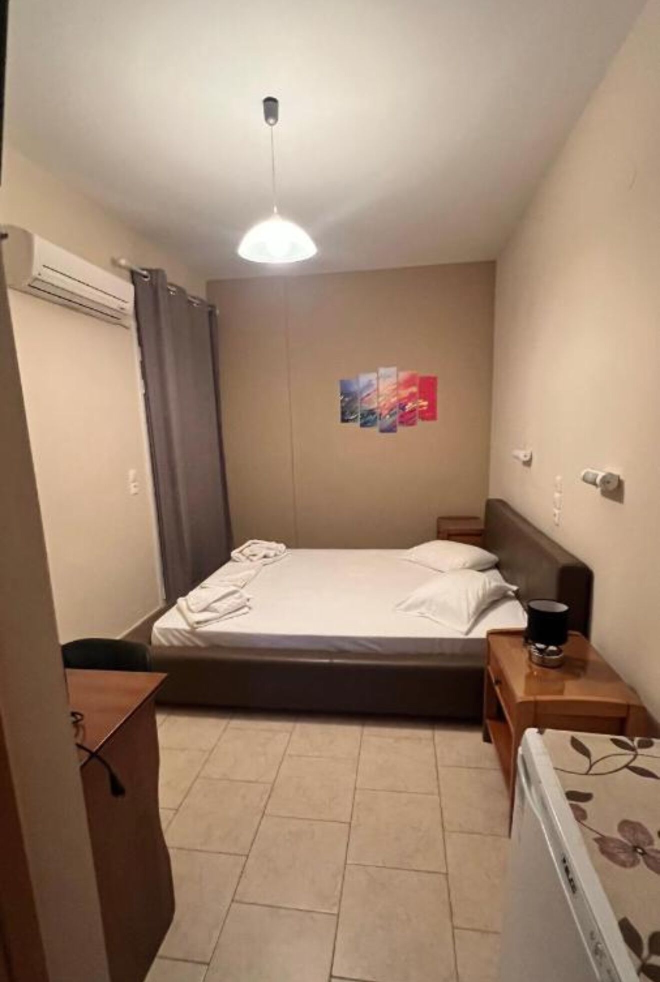 Een kleine, netjes ingerichte slaapkamer met een enkel bed, een nachtkastje, een airconditioning en een wandlamp.