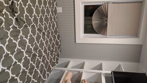 A bedroom with a patterned bedspread, a window with blinds, a white bedside table, and a TV.