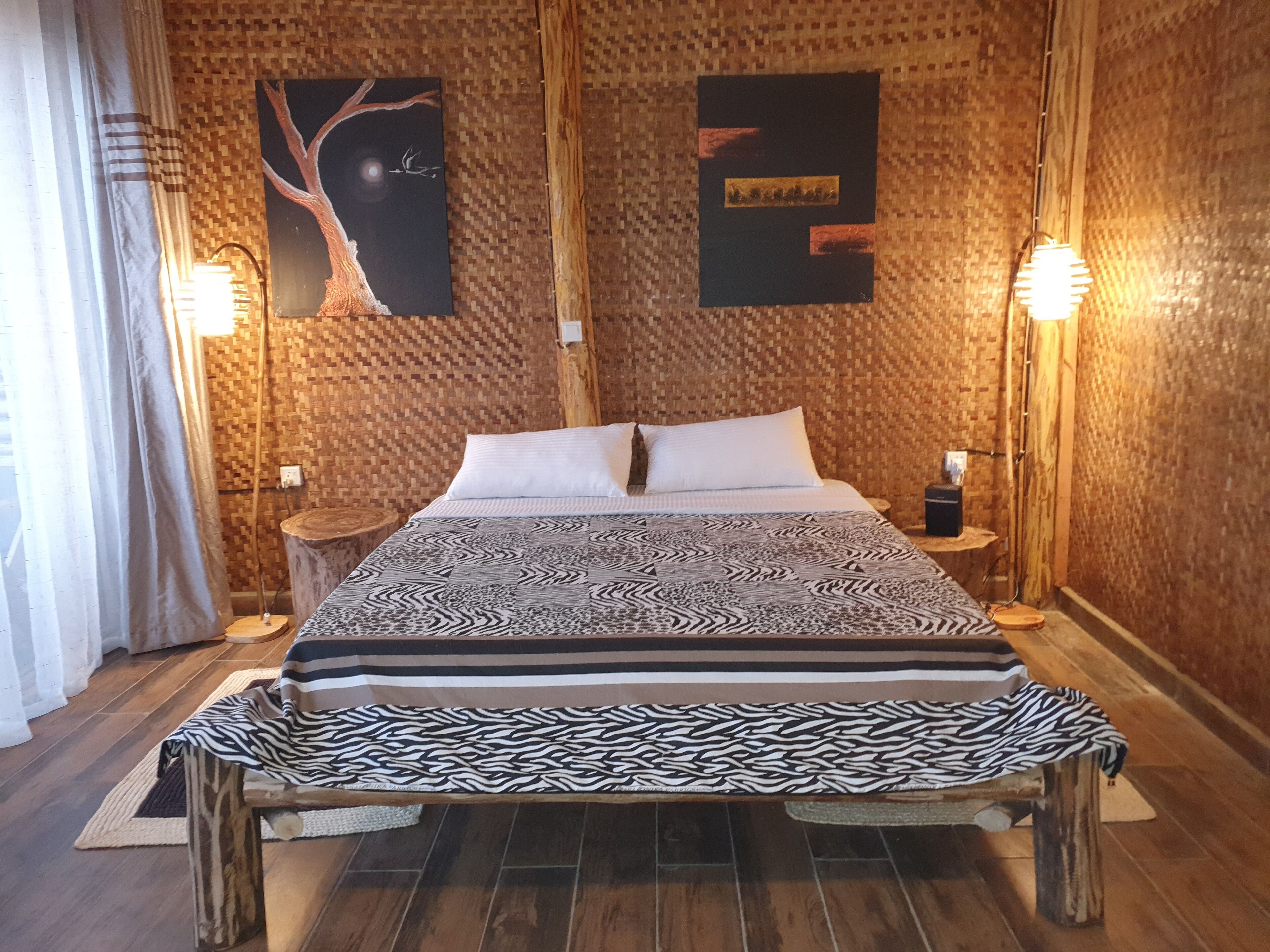 A bedroom with a patterned bedspread, two wall-mounted artworks, and a bench at the foot of the bed.