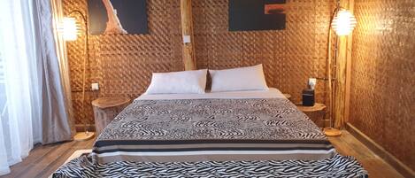 A bedroom with a patterned bedspread, two wall-mounted artworks, and a bench at the foot of the bed.