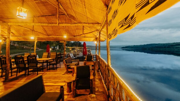 An outdoor dining area with wooden tables and chairs, a thatched roof, and a view of a calm body of water. - Vintage Cottage (Gishari)