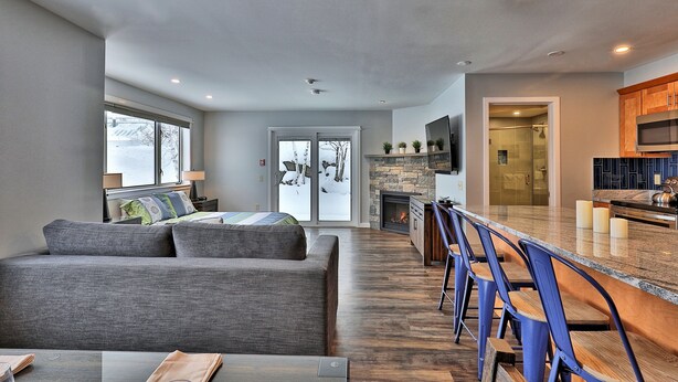 Ein modernes Wohnzimmer mit grauem Sofa, einem Essbereich mit blauen Stühlen, einer Küche mit dunklen Schränken und einem Kamin aus Stein.