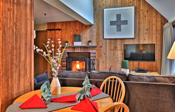 A cozy living room with a fireplace, wooden walls, a TV, and a dining area with a vase of flowers.
