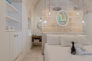 Une chambre avec un mur en pierre, un miroir rond, un lit avec du linge de lit blanc et des luminaires suspendus.