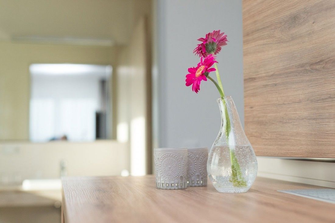 En genomskinlig glasvas med rosa blommor står på ett träbord bredvid ett dekorativt glashållare för ljus.
