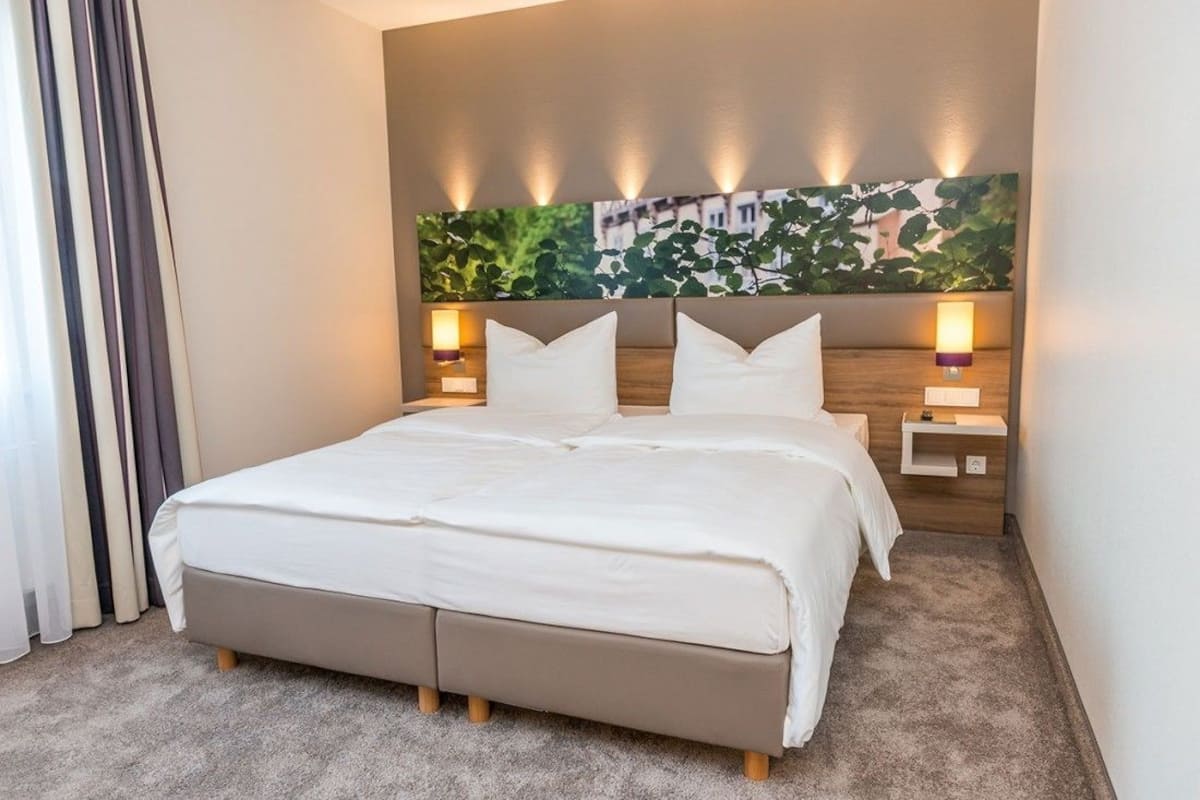 Habitación de hotel con una cama grande, dos mesitas de noche con lámparas, un cabecero con una obra de arte de motivos vegetales, y vistas a edificios a través de la ventana.