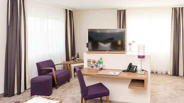 A hotel room with a flat-screen TV, a desk with a phone, a chair, a small table with a basket, and a sofa. - Stratmanns Hotel (Lohne)