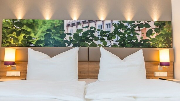 Habitación de hotel con una cama grande, dos lámparas de noche y un cuadro de follaje colgado en la pared.