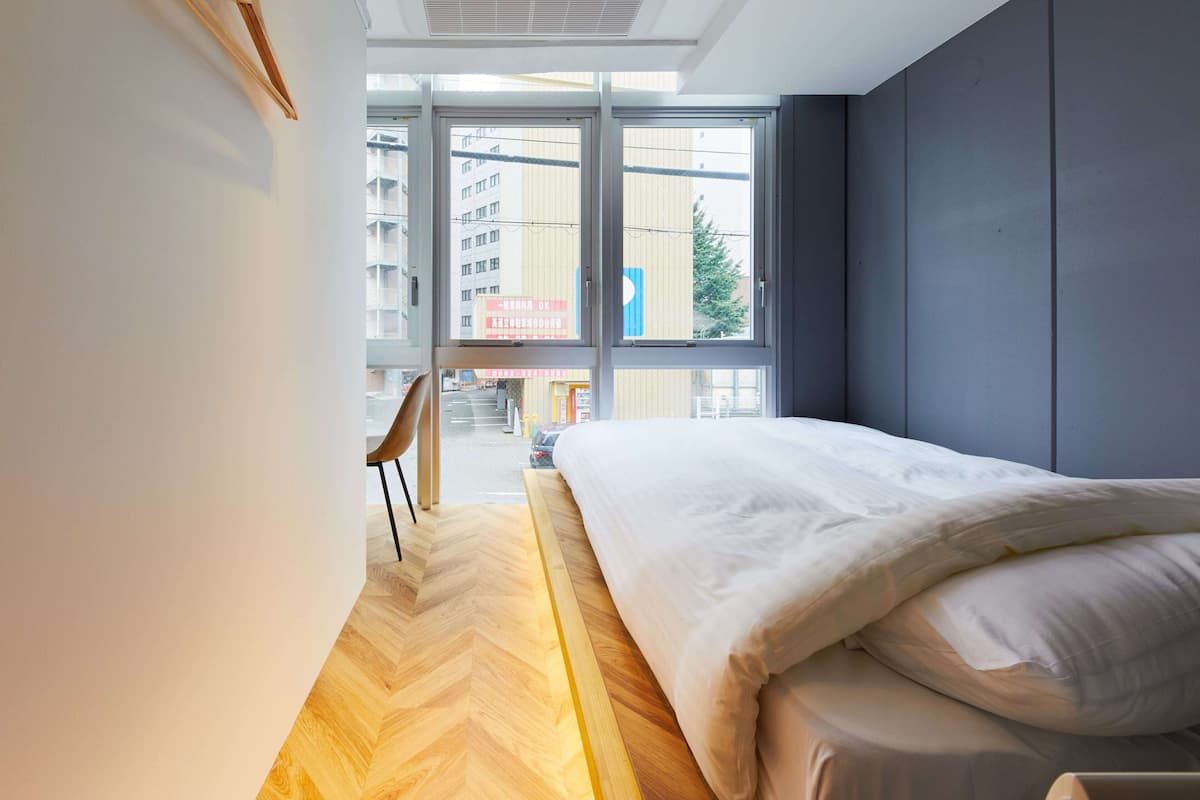 A modern hotel room with a large bed, a wooden floor, and a view of the city through large windows.